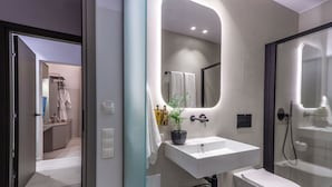 Une salle de bain moderne avec un lavabo blanc, des toilettes et une douche à parois de verre. Un miroir à la forme originale est placé au-dessus du lavabo, et une plante en pot se trouve sur le comptoir.