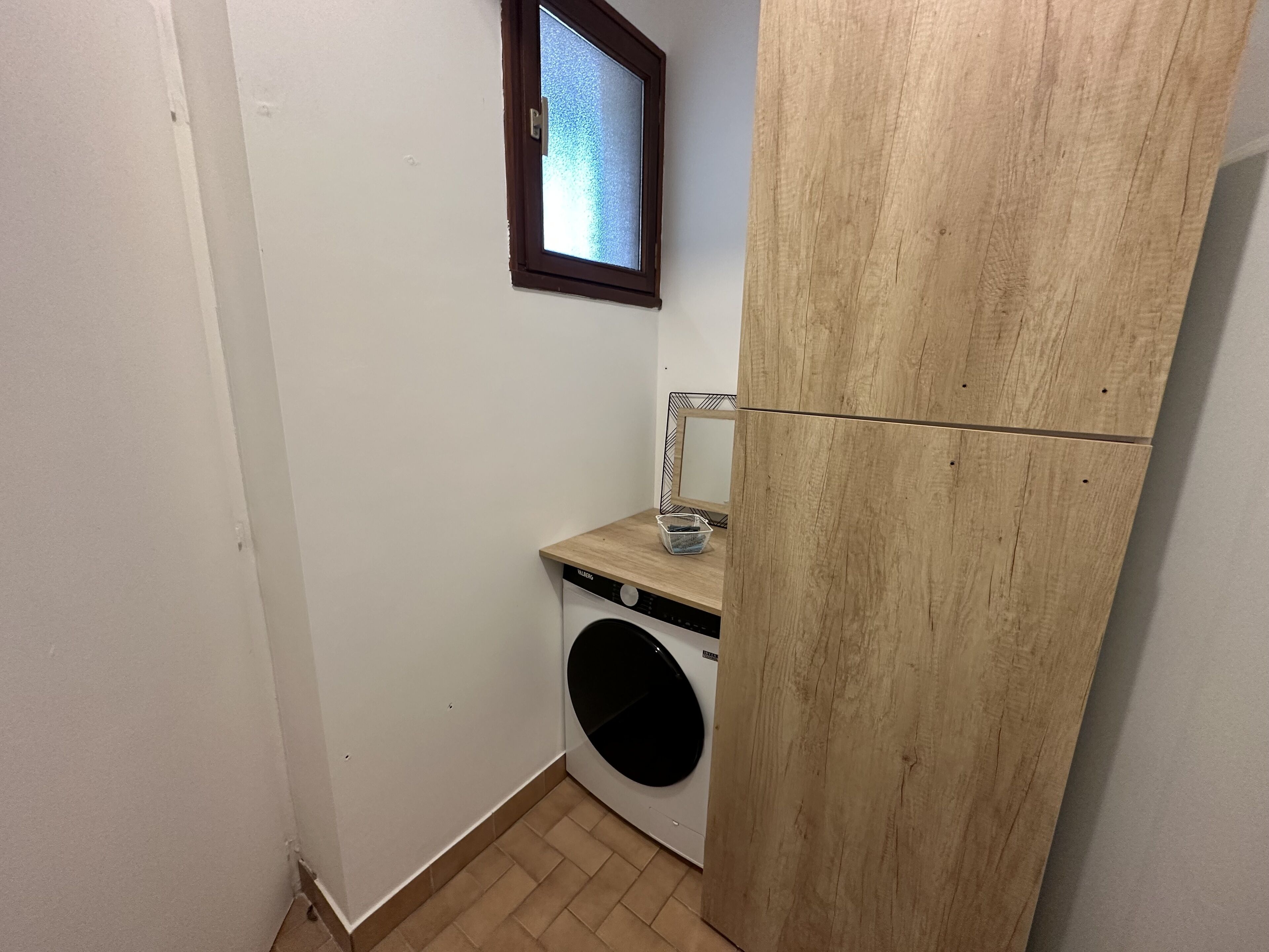 A laundry room with a washing machine, a small wooden cabinet, and a framed picture on the wall.