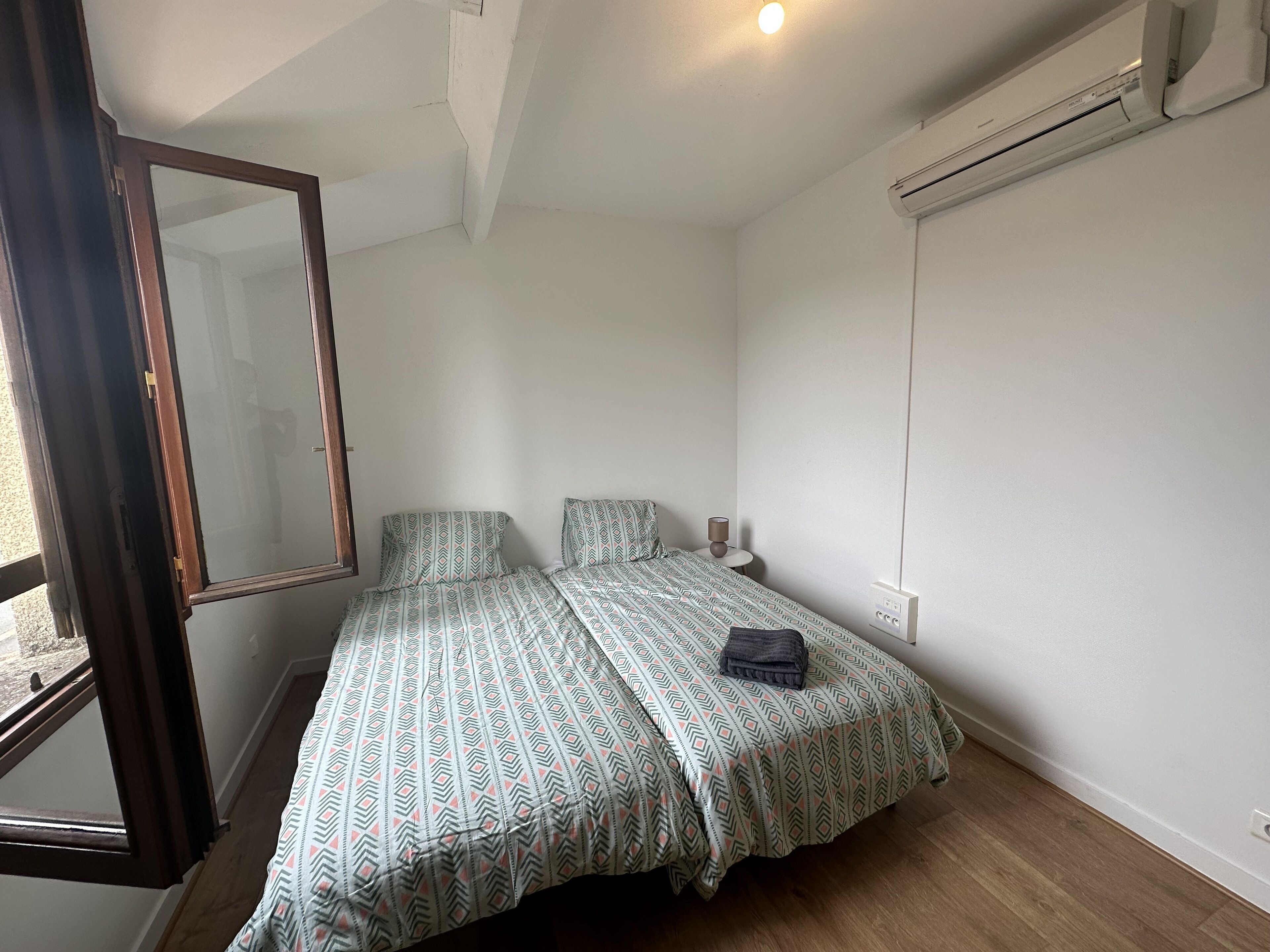 Ein kleines, ordentlich eingerichtetes Schlafzimmer mit Bett, Nachttisch, Spiegel und einer Klimaanlage.