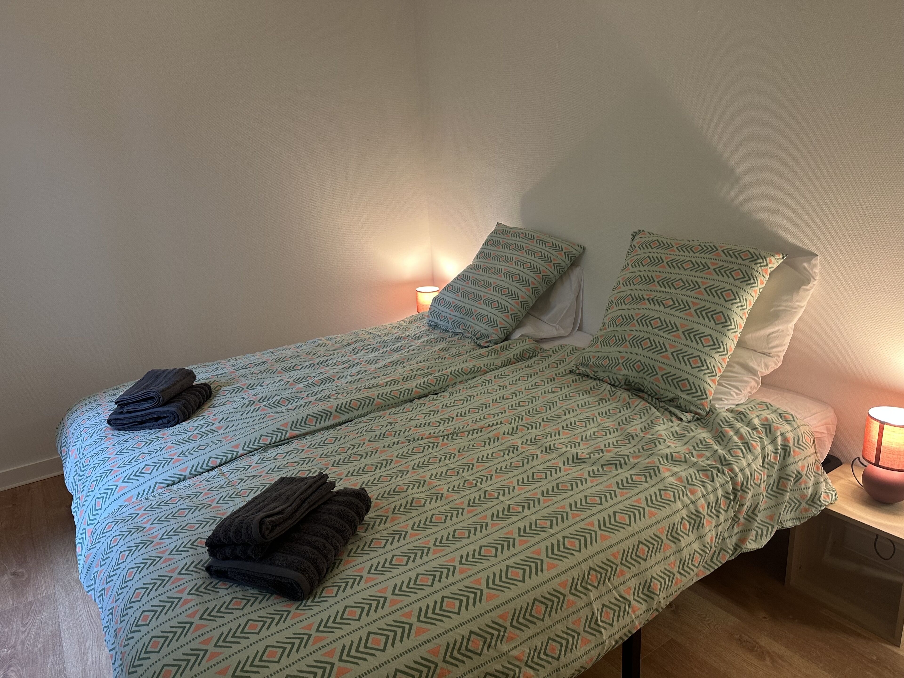 Een enkel bed met beddengoed met dessin, twee kussens en een opgevouwen handdoek. Aan de rechterkant staat een bedlamp.