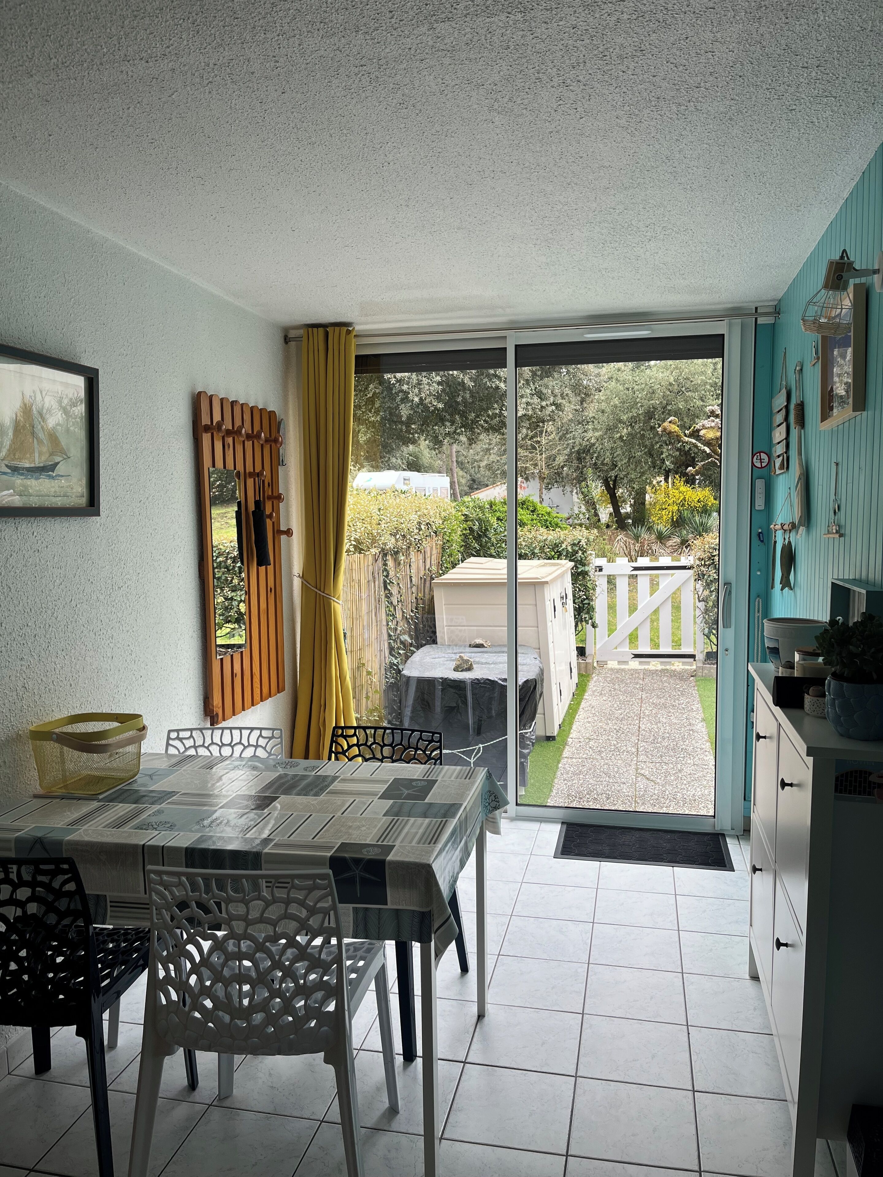 A dining area with a table and chairs, a window with yellow curtains, and a view of a garden and white fence outside.