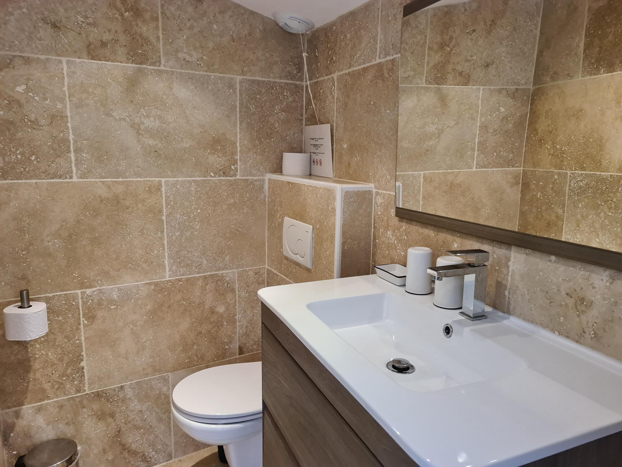 Ein Badezimmer mit beigen Fliesen, einem weißen Waschbecken, einem Spiegel und einer Toilette.