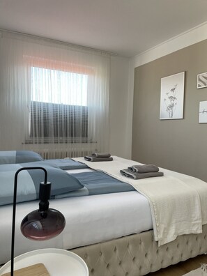 A bedroom with a bed, a bedside table, a lamp, a window with sheer curtains, and wall art. - GREEN HILL Apartment (Bremerhaven)