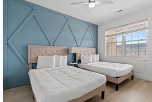 Une chambre avec deux lits, un mur d’accent bleu, un ventilateur de plafond et une fenêtre avec des stores.
