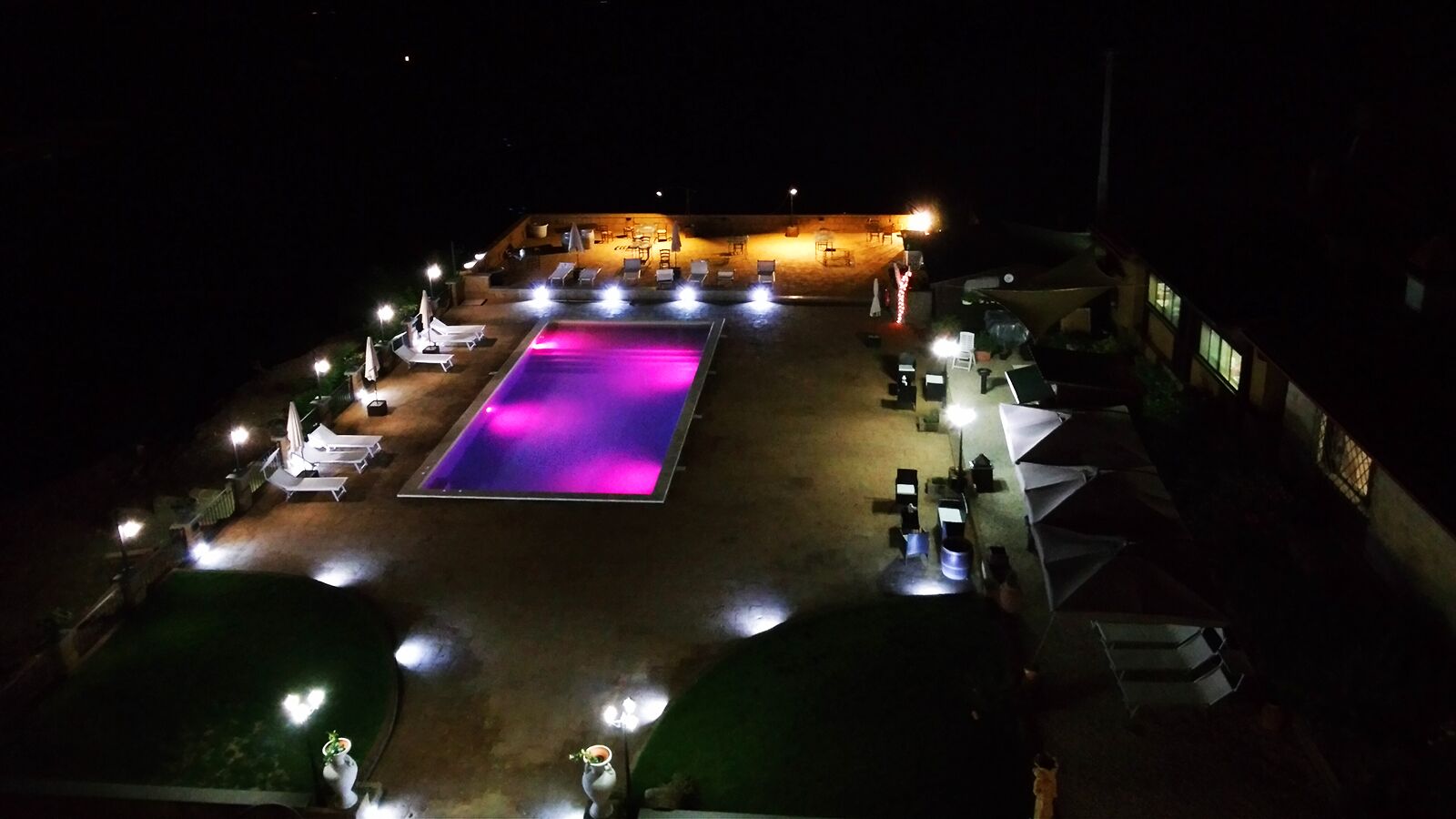 Vue nocturne d’un espace piscine avec des chaises longues et des parasols, entouré d’immeubles.