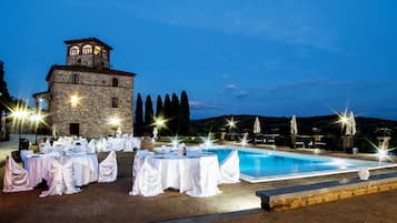 Un espace de restauration aménagé au bord de la piscine, avec des tables dressées pour le repas, un bâtiment en pierre doté d’une tour et un ciel nocturne dégagé.