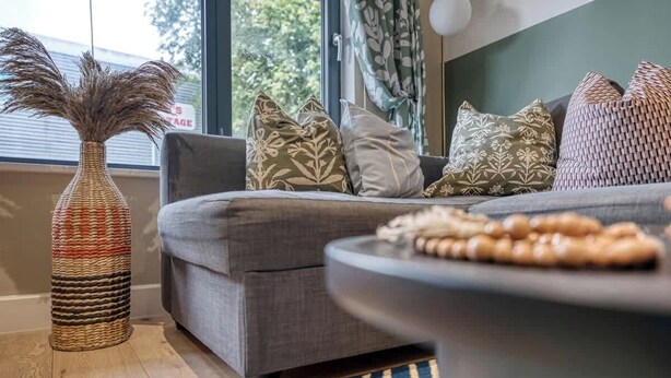 Ein Wohnzimmer mit grauem Sofa, gemusterten Kissen, einer gewebten Vase und einem runden Tisch mit Holzperlen.