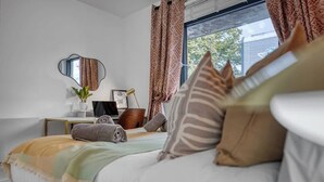 Ein Schlafzimmer mit Bett, Kissen, einem Nachttisch mit Lampe, einem Spiegel und einem Fenster mit Vorhängen.