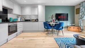 Eine moderne Küche mit weißen Schränken, ein Essbereich mit blauen Stühlen, einem Flachbildfernseher und einem Koffer.