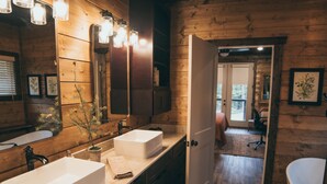 A wooden cabin bathroom with two sinks, a large mirror, and a door leading to a bedroom. - Experience the most stunning views the Ozarks have to offer (Jasper)