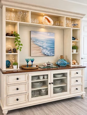 A white cabinet with glass-fronted shelves holding plates and glasses, a painting of a beach, and decorative items.
