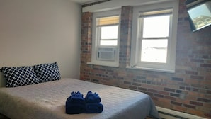 Ein Schlafzimmer mit einem Bett, zwei Kissen, einem Fenster mit Jalousien und einer Ziegelwand.