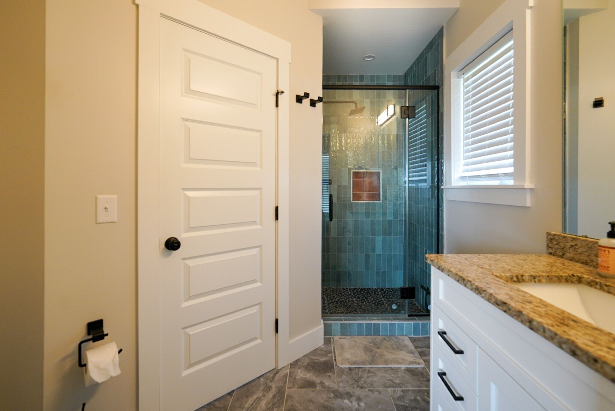A bathroom with a white door, a shower with blue tiles, and a granite countertop.