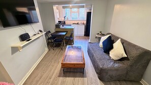 A compact living space with a sofa, a coffee table, a TV mounted on the wall, a dining area with chairs, and a kitchen in the background. - London Apartment - Near King’s Cross St Pancreas! (Greater London)