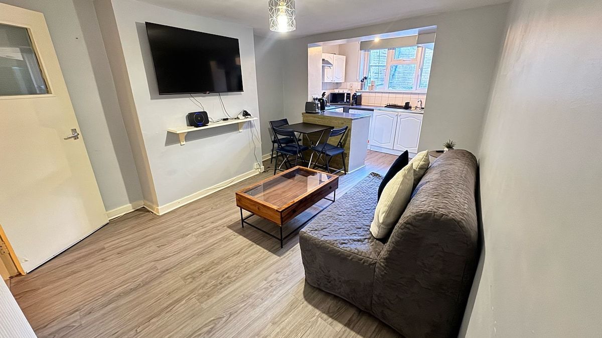 Un espacio habitable compacto con un sofá, una mesa de centro, un televisor montado en la pared y una cocina visible a través de una puerta abierta.