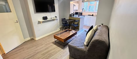 Un espacio habitable compacto con un sofá, una mesa de centro, un televisor montado en la pared y una cocina visible a través de una puerta abierta.