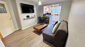 A compact living space with a sofa, a coffee table, a TV mounted on the wall, and a kitchen area visible through an open door. - London Apartment - Near King’s Cross St Pancreas! (Greater London)