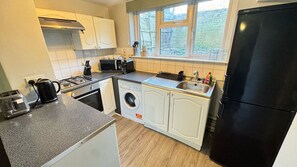 A compact kitchen with a gas stove, a washing machine, a refrigerator, and a window with a view of greenery. - London Apartment - Near King’s Cross St Pancreas! (Greater London)