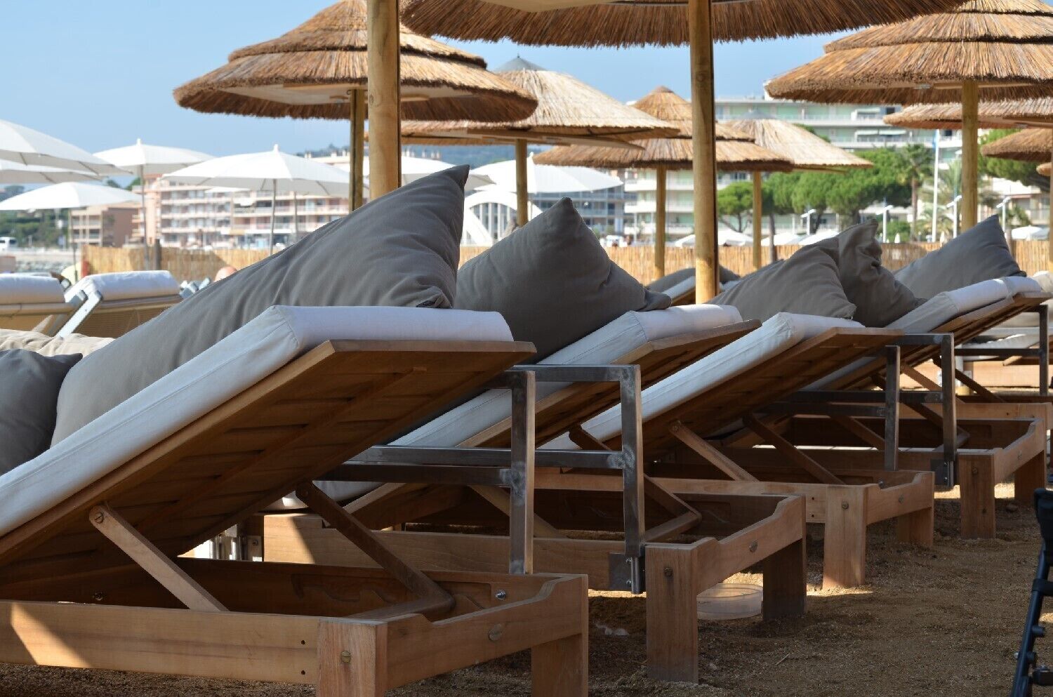 Una hilera de tumbonas de madera con cojines grises, dispuestas en una playa arenosa con sombrillas de paja al fondo.