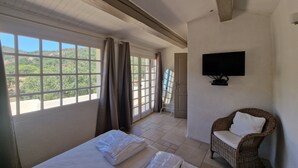 A room with a bed, a wicker chair, a TV, and a view of greenery through large windows. - Heerlijk Vakantiehuis Les Pennes 4 met 3 Slaapkamers, Zeer Groot Privé Terras (Le Plan-de-la-Tour)