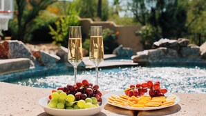 Une table avec deux coupes de champagne, un bol de raisins et une assiette de fromage et de crackers, près d’une piscine.