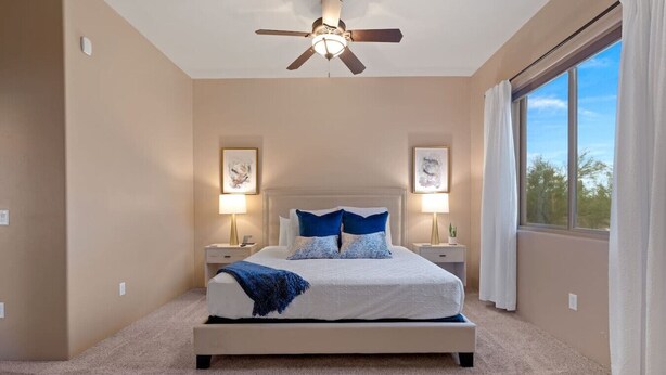 Une chambre à coucher comprenant un lit, deux tables de chevet, un ventilateur de plafond et une grande fenêtre avec des rideaux.