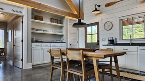 Eine moderne Küche mit weißen Schränken, einem Holzespeisetisch und Stühlen. Regale mit Geschirr und Gläsern, eine Uhr und ein Fenster mit Jalousien sind ebenfalls vorhanden.
