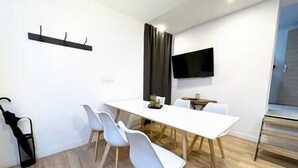 Un área de comedor moderna con una mesa blanca, cuatro sillas blancas, un televisor de pantalla plana montado en la pared y suelo de madera.