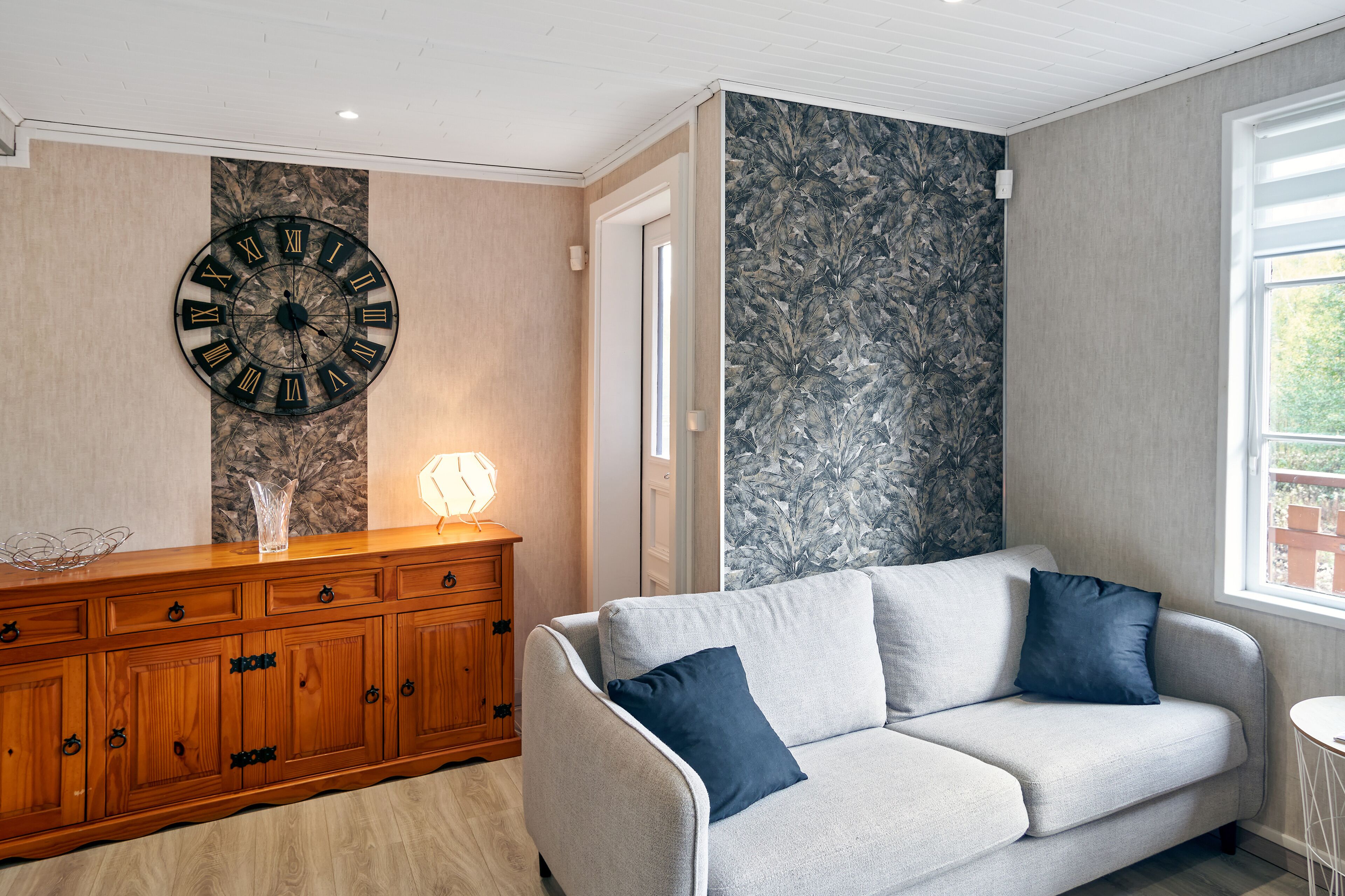 Ein Wohnzimmer mit einem Holzkabinett, einem grauen Sofa mit blauen Kissen, einer großen Wanduhr und einer Wand mit gemustertem Tapetenpapier.