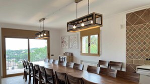 A dining area with a long wooden table and chairs, a hanging light fixture, and a view of greenery outside. - Villa Cabañas with infinity pool, gardens and barbecue in Calonge, Costa Brava (Calonge)