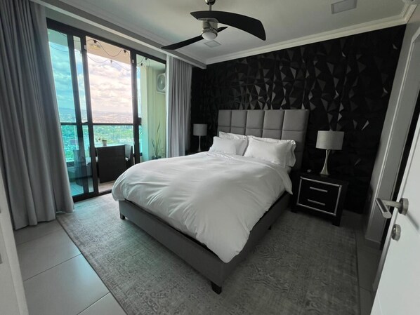 A modern bedroom with a large bed, a ceiling fan, a nightstand, and a view through a glass door to an outdoor area.