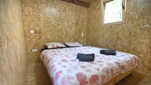 A small, single-bed room with a wooden floor and walls, a window with a view of greenery, and a bed with a floral bedspread and pillows. - TreeHouses Ljubač Glamping Robinson (Razanac)
