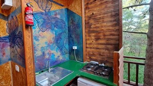 A small, rustic kitchen with a green countertop, a sink, and a stove. A fire extinguisher is mounted on the wall, and there is a window overlooking a forested area. - TreeHouses Ljubač Glamping Robinson (Razanac)