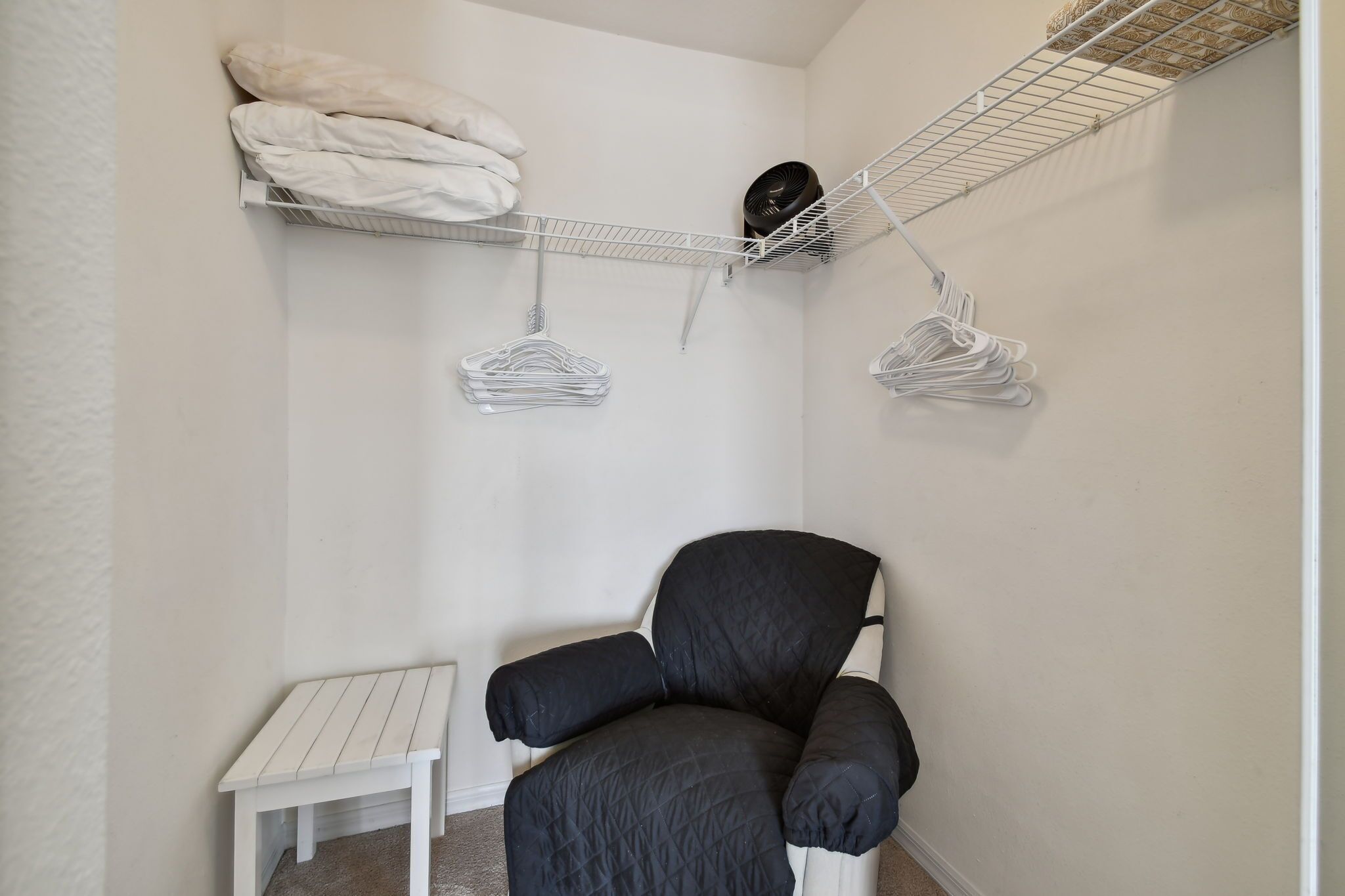 Habitació petita i ben ordenada amb una cadira negra, un taulell blanc i una prestatgeria metàl·lica amb roba doblegada i un ventilador.