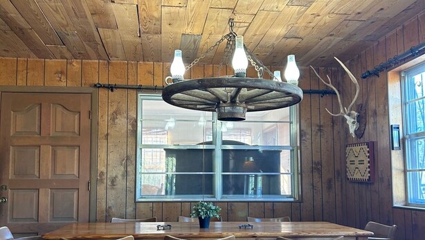 Un intérieur rustique avec des éléments en bois, notamment une grande table et des chaises en bois, un lustre orné d’ampoules et un crâne de cerf accroché au mur.