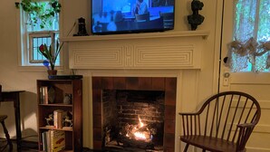Ein gemütliches Wohnzimmer mit Kamin, einem darüber montierten Fernseher, einem Bücherregal und einem bequemen Sessel.