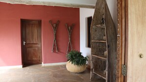 Una puerta de madera, dos esculturas fijadas a la pared, una planta grande en maceta y una escalera de madera rústica.