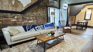 A cozy living room with a brick wall, a sofa with a 'Penn State' pillow, a coffee table, and a dining area with a table and chairs. - Stylish, Historic PSU-themed Apt.—2 blocks from Waterfront (Bellefonte)