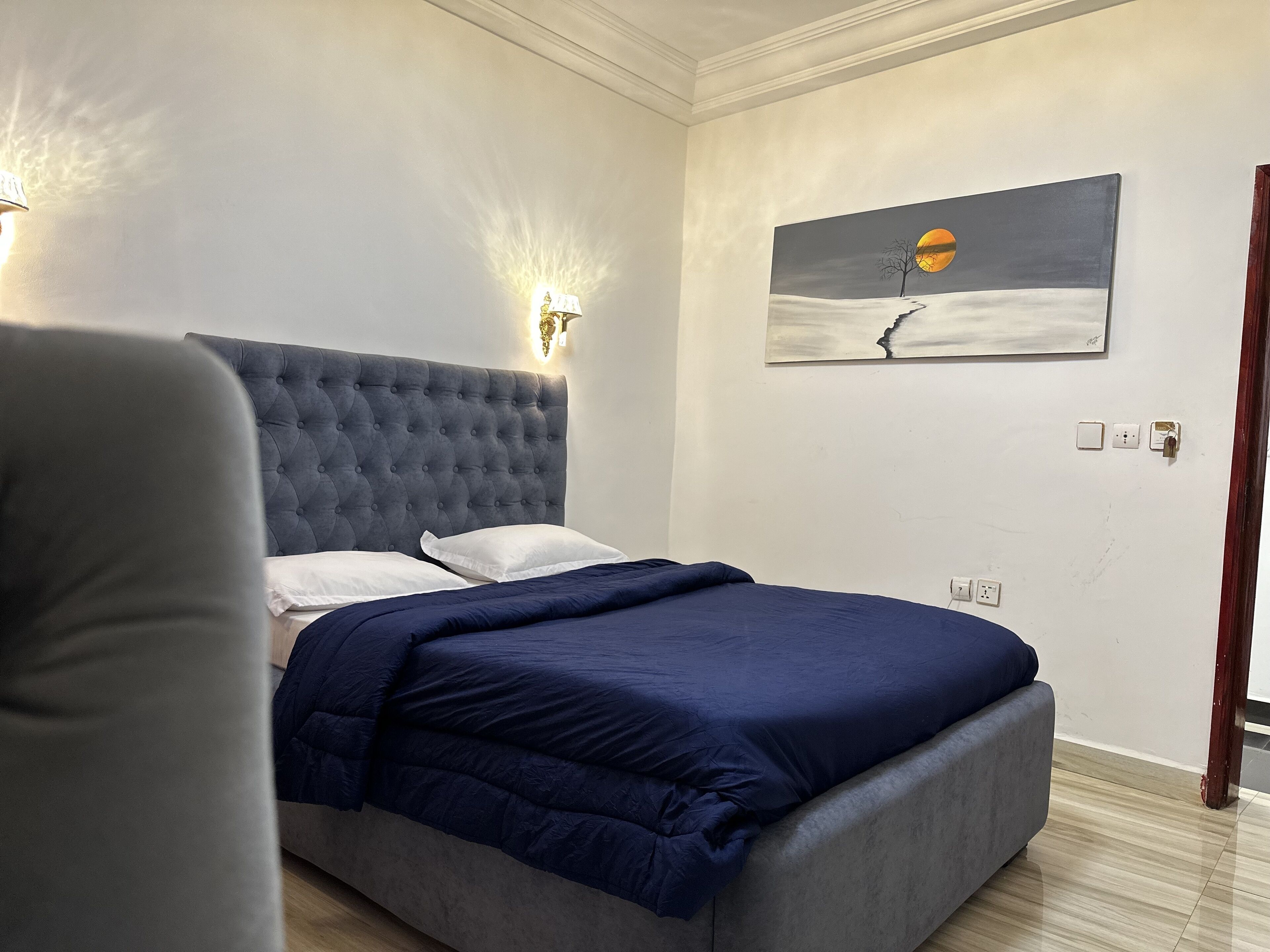 A bedroom with a tufted headboard, a bed with a dark blue bedspread, a painting of a sunset on the wall, and a nightstand with a lamp.