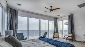 A spacious bedroom with a large bed, two chairs, a ceiling fan, and a view of the sea through large windows. - Stunning Oceanfront 5 Bed House with Infinity Pool (West Bay)