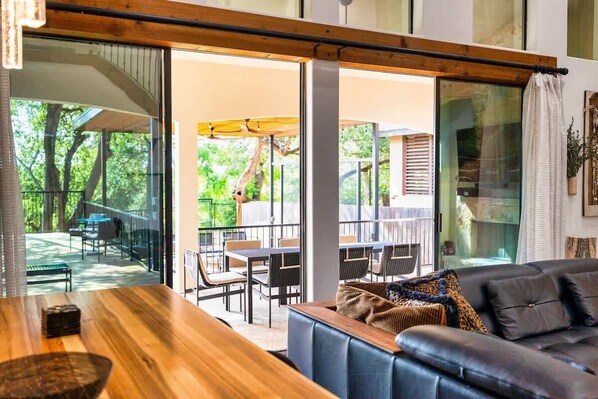 A modern living room with a wooden table and a leather sofa, large windows with a view of an outdoor patio area.