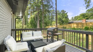 Eine überdachte Terrasse im Freien mit Korbmöbeln, einem Grill und Blick auf ein bewaldetes Gebiet.