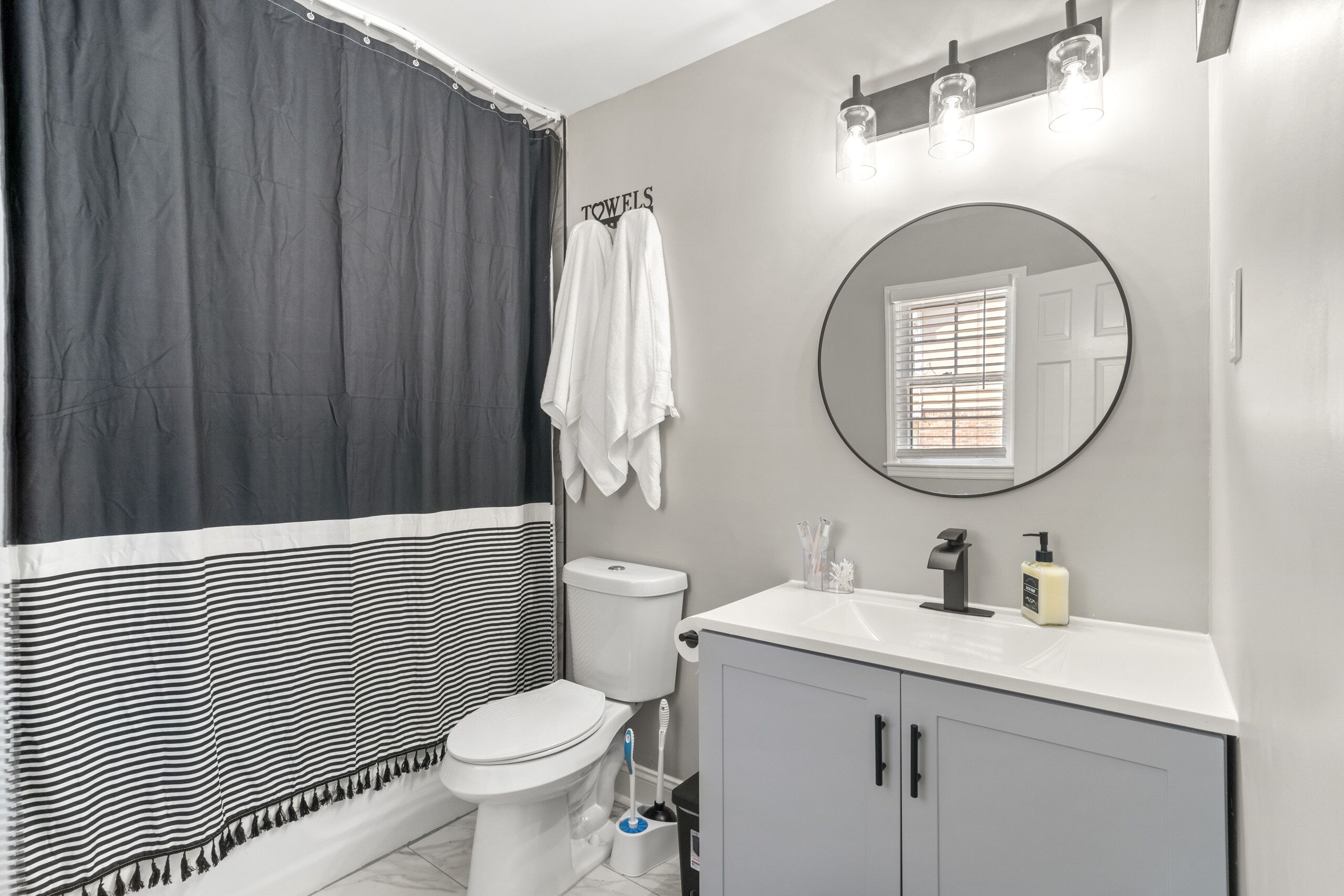 Un baño con inodoro, lavabo y espejo. Se ve un perchero con toallas colgadas.