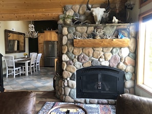 Una chimenea de piedra con cuernos y cráneos, un comedor con mesa y sillas de madera, y una cocina con refrigerador y alacenas.