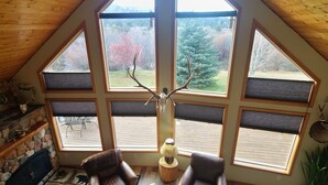 A cozy loft bedroom with a sloped ceiling, large windows offering a view of the outdoors, a ceiling fan, and a stone fireplace. - Three bedroom,  two bath on the Middle Fork of Rock Creek (Philipsburg)