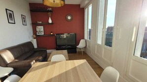 Un soggiorno moderno con un tavolo da pranzo in legno, sedie bianche, un divano in pelle marrone, una TV e una parete di accento rossa con orologio e oggetti decorativi.