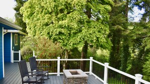 En terrasse med en ildsted i sten og to gyngestole, omgivet af et hvidt gelænder og frodig beplantning.