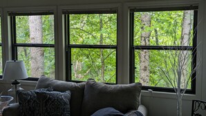 Ein Zimmer mit einer Couch, einer Lampe und drei Fenstern mit Blick auf einen Wald.