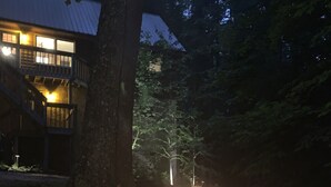 Un fuoco da campo acceso in una fossa in metallo, circondato dall'erba, con una casa sullo sfondo.