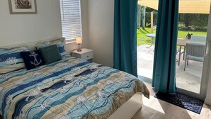 Une chambre à coucher comprenant un lit, une table de chevet, un ventilateur de plafond, une fenêtre avec des stores et une vue sur une terrasse extérieure.